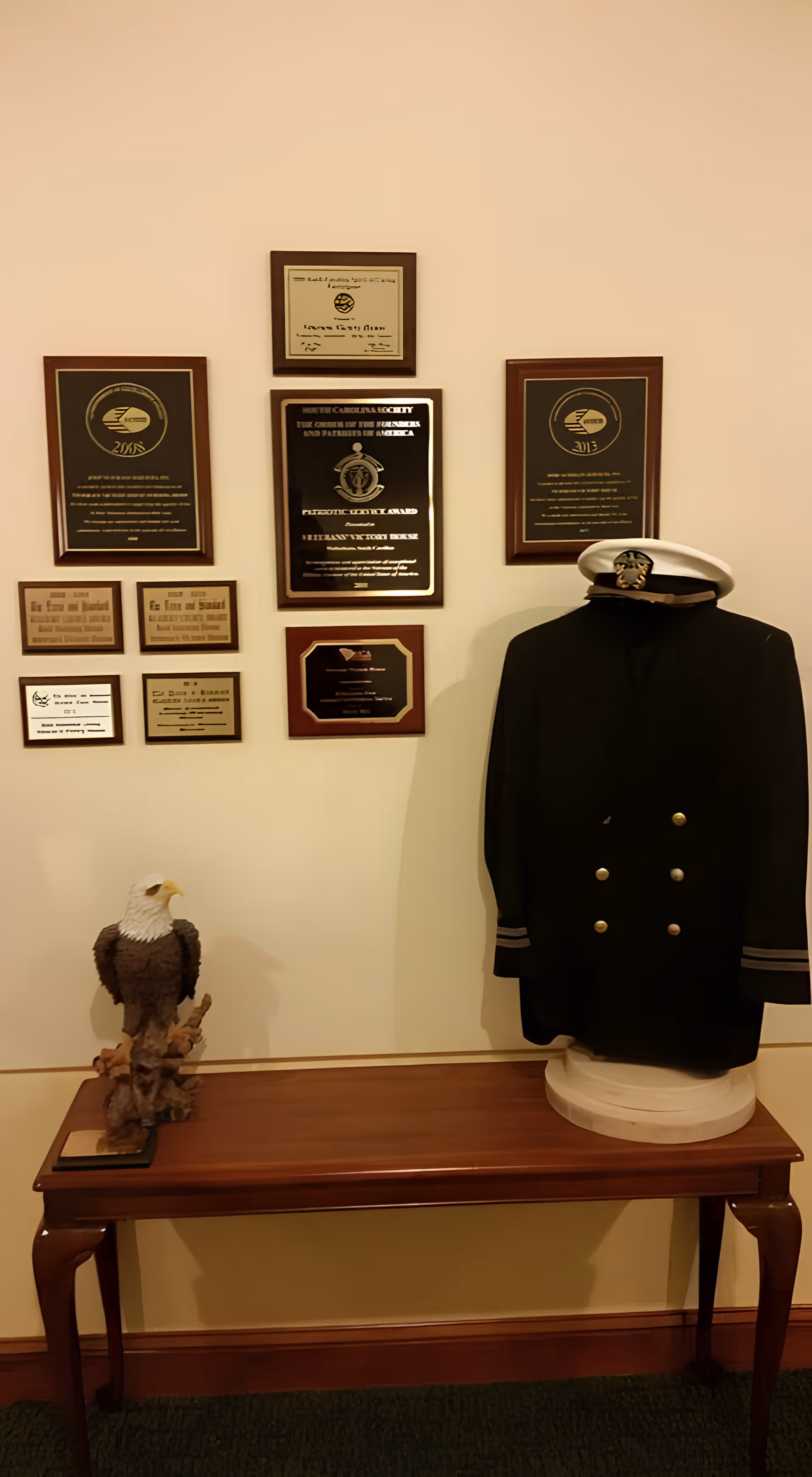 A display wall featuring several plaques and awards mounted on a beige wall above a wooden table. On the table, there is a sculpture of a bald eagle and a mannequin torso dressed in a dark military uniform jacket with gold buttons and a white officer's hat.