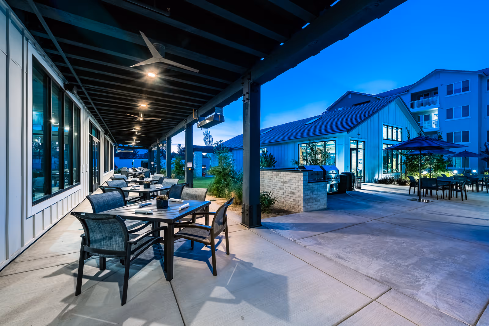 Outdoor patio area at dusk with multiple tables and chairs arranged under a covered walkway and open sky. There are ceiling fans and lights under the covered area. In the background, there is a building with large windows and an outdoor grill station, along with more seating and umbrellas.