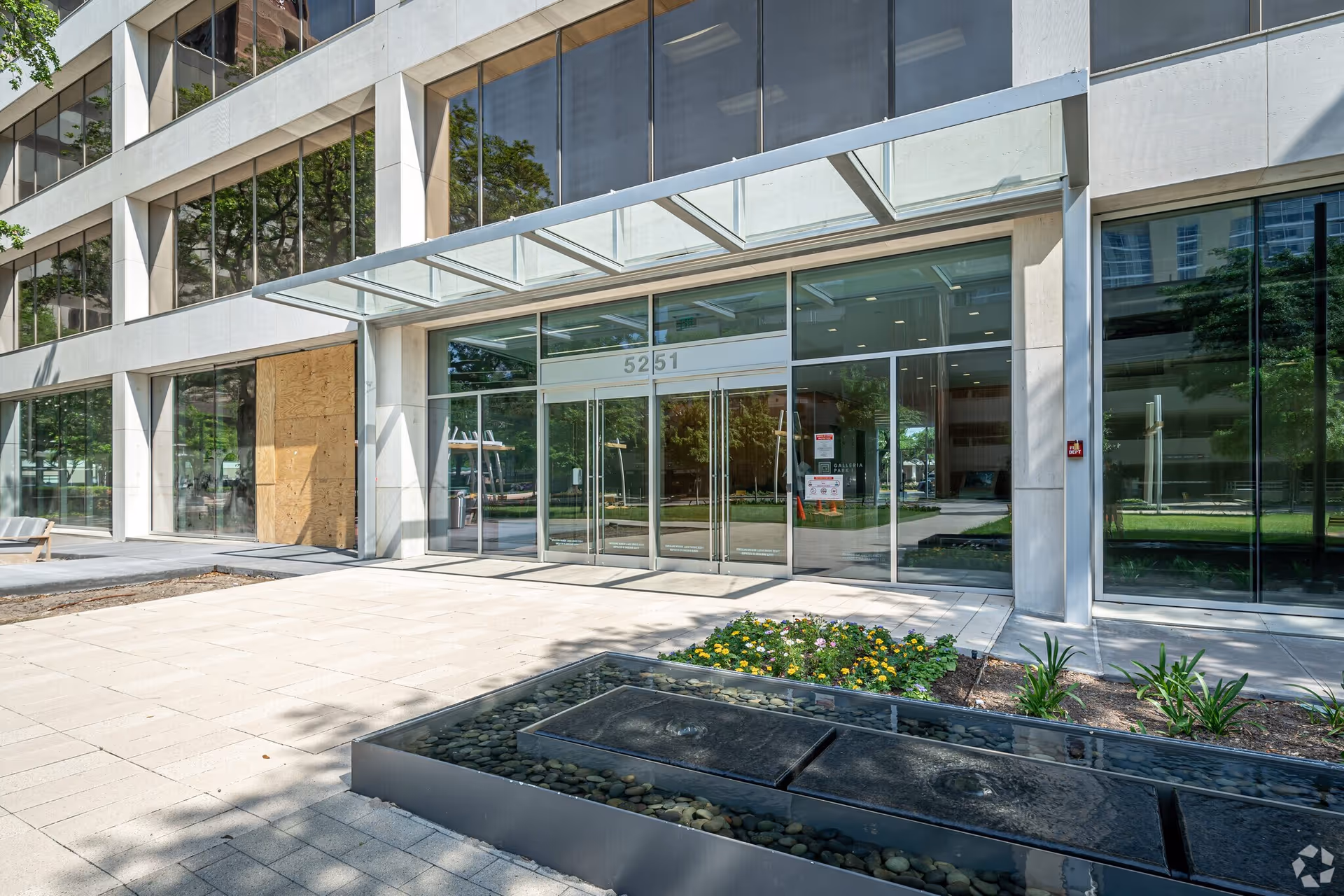 Modern glass-front entrance of a large building with landscaping and water feature.