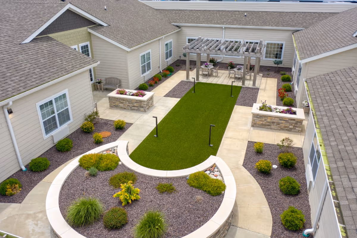 A landscaped outdoor courtyard area at an assisted living facility featuring a green lawn, stone planters with flowers and shrubs, a curved garden bed with various plants, concrete walkways, and a pergola with tables and chairs underneath. The courtyard is surrounded by beige buildings with multiple windows.