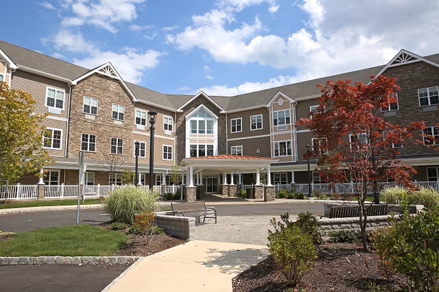 Front exterior view of a senior living facility building with three stories, featuring a covered entrance, multiple windows, landscaped garden areas with shrubs and trees, benches, and a clear blue sky with some clouds.