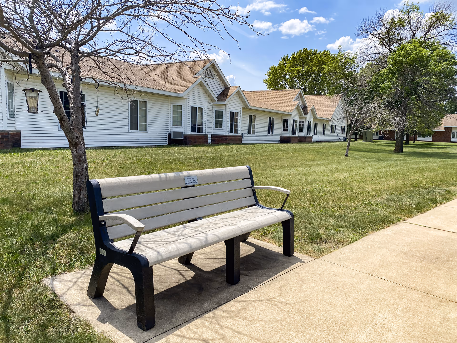 A bench on a sidewalk beside a green lawn and white cottages under blue sky.