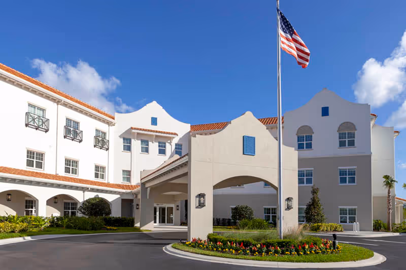 Front entrance of a Mediterranean-style senior living building with an arched porte-cochère, landscaped circular driveway, and an American flag.