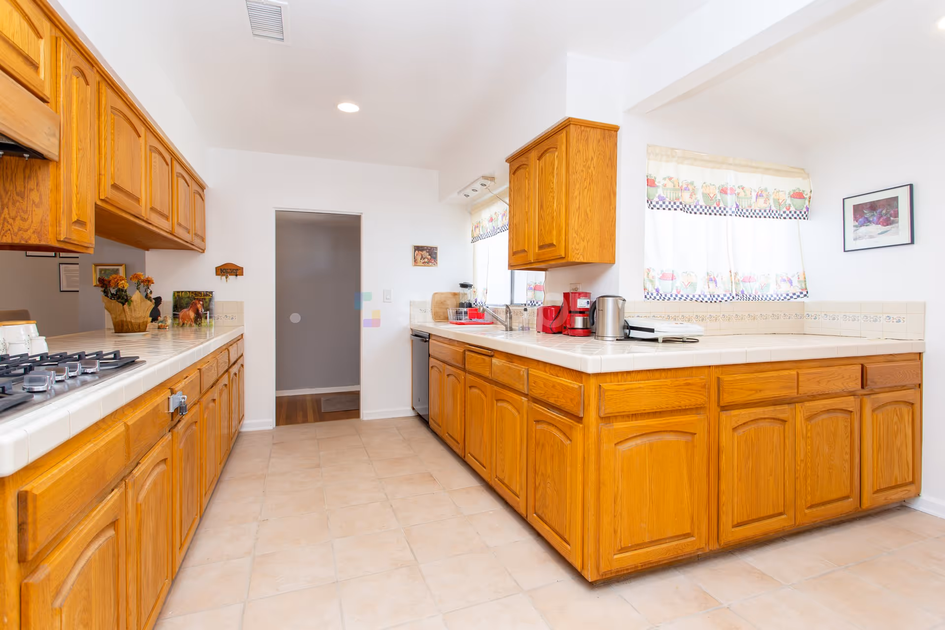 Bright kitchen with wooden cabinets and tiled countertops. The kitchen features a gas stove on the left side, a sink under a window with decorative curtains, and various small appliances including a red coffee maker and a kettle. The floor is tiled, and the walls are painted white with a framed picture hanging on the right side.