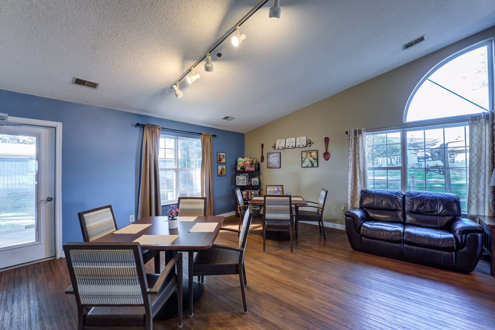 Bright shared dining and living area with tables, chairs, and a leather sofa.