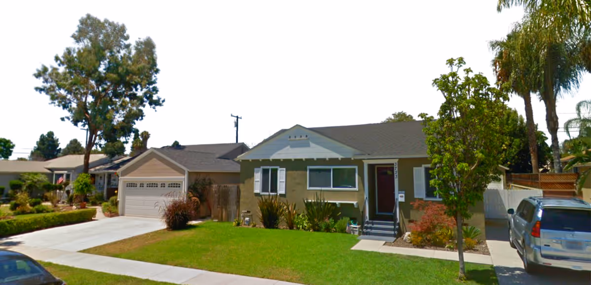 Front exterior view of a single-story residential building with a green lawn, trees, and a driveway with a parked car.