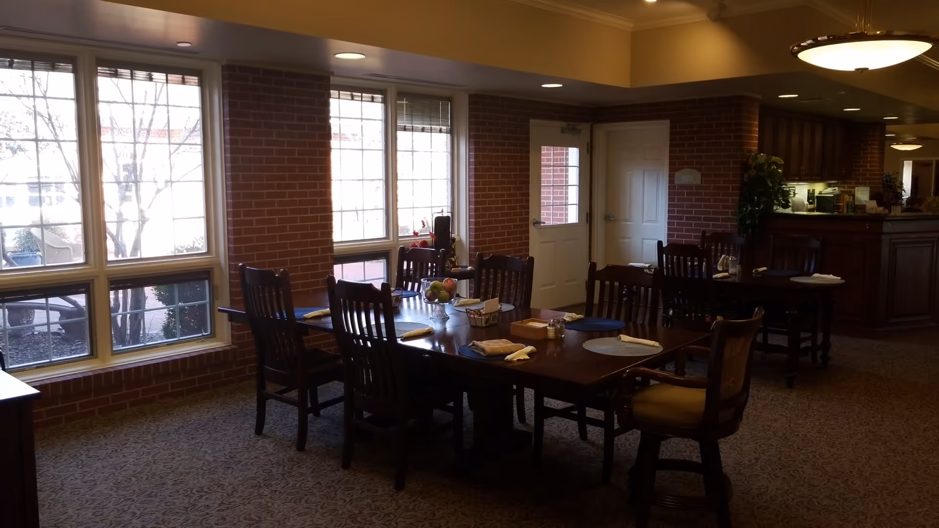 Spacious dining room with a long wooden table, chairs, brick columns, and large windows.
