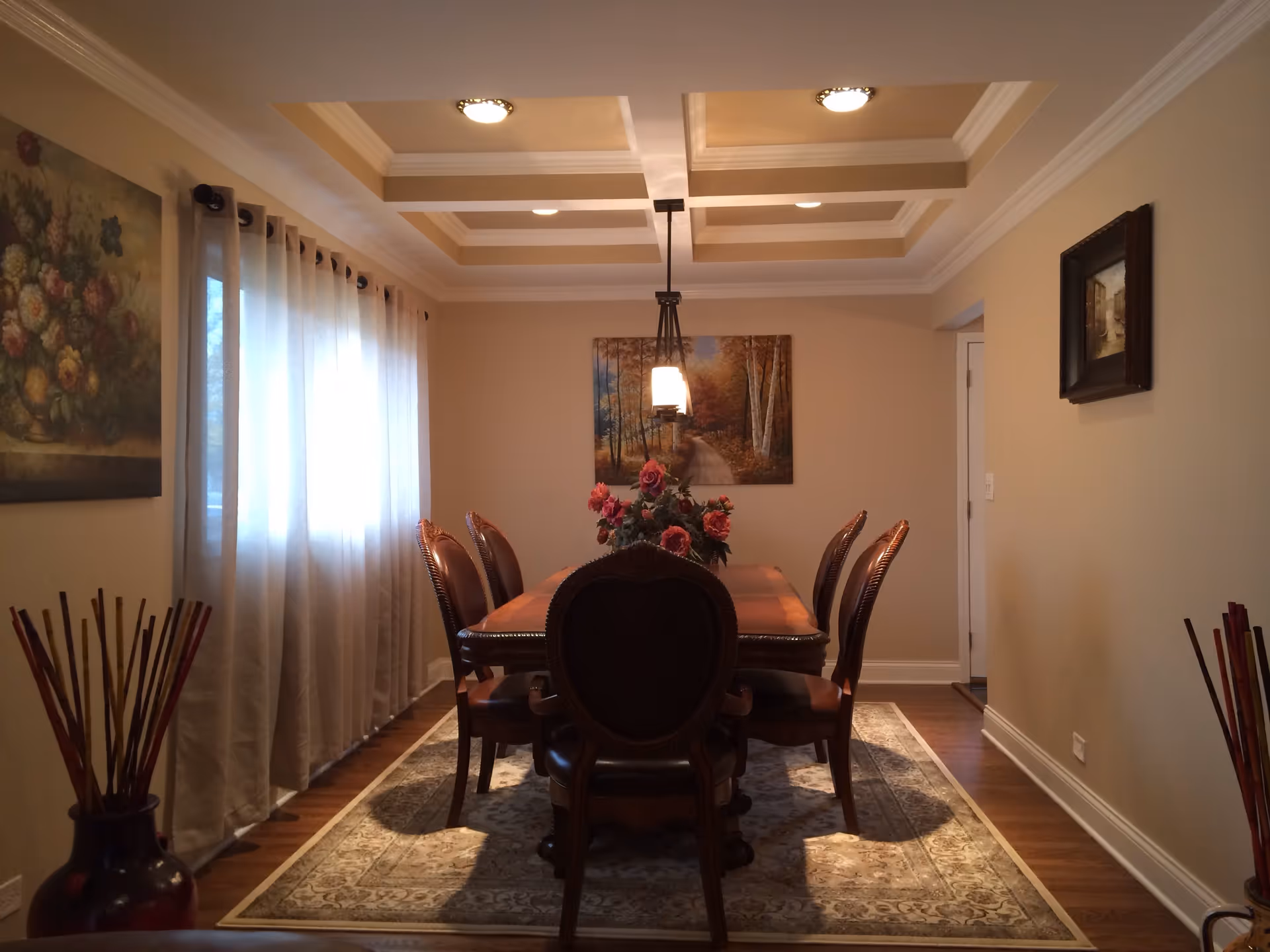 A formal dining room with a wooden dining table and six matching chairs. The table has a floral centerpiece with pink flowers. The room features a coffered ceiling with recessed lighting and a hanging light fixture above the table. There are three paintings on the walls and sheer curtains covering the window on the left side. The floor has a patterned area rug under the table and hardwood flooring around it.