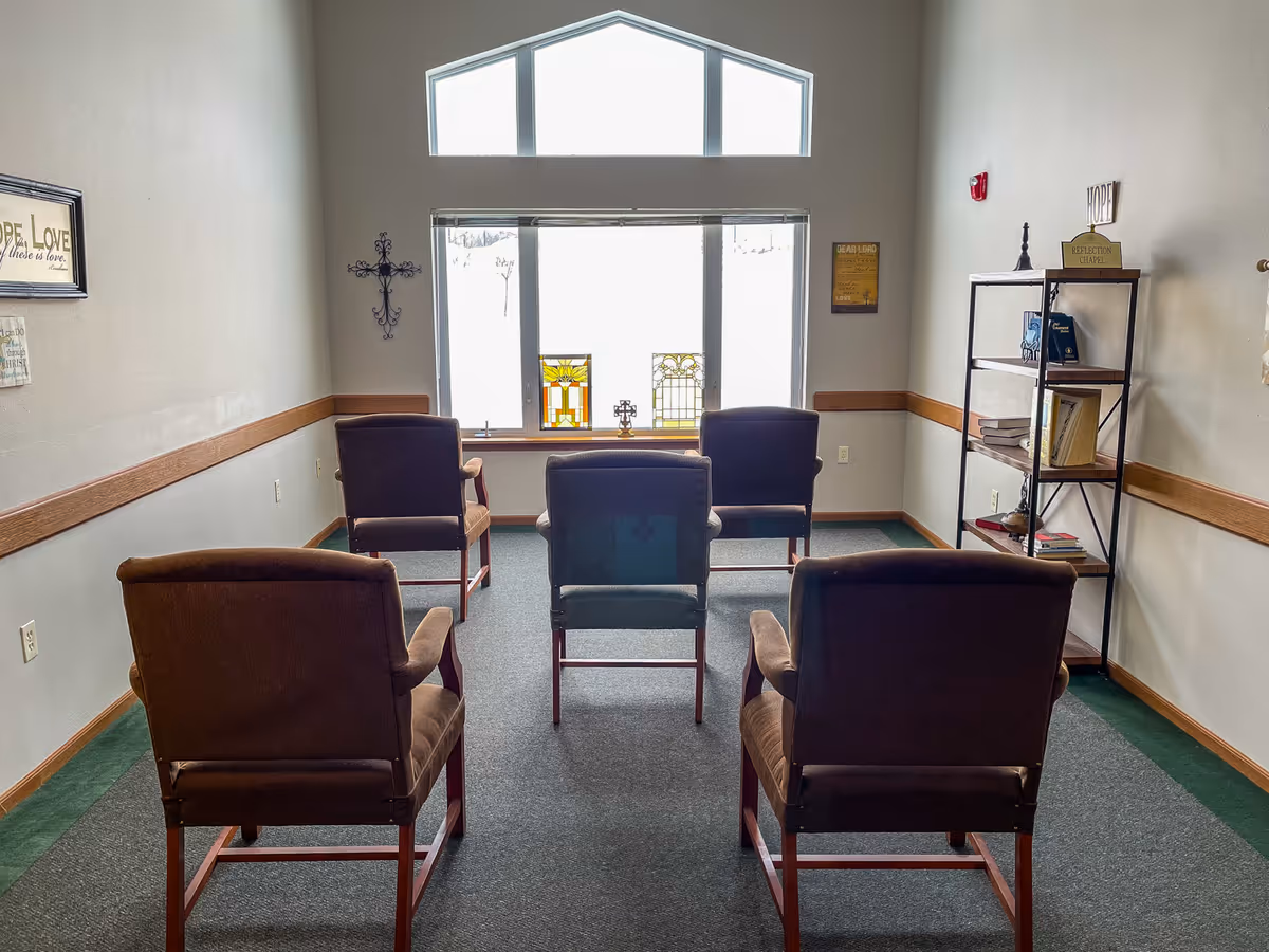 Rows of brown cushioned chairs face a windowed wall with a cross and shelves.