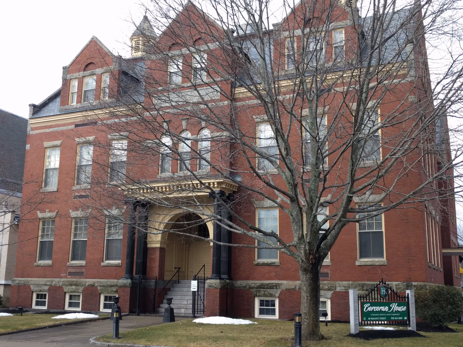 Brick multi-story building with entrance steps and a large tree in front; sign.