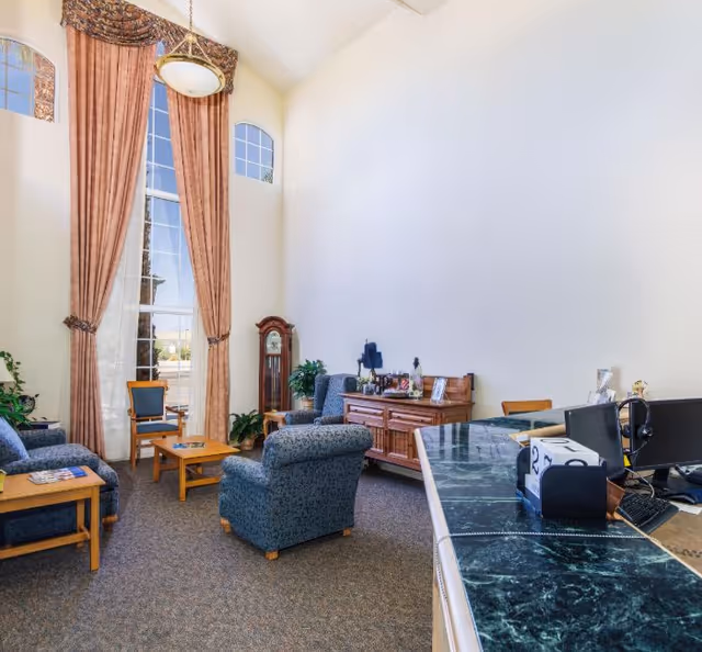 Spacious senior living lounge with blue upholstered chairs, tall windows, curtains, and a marble counter.