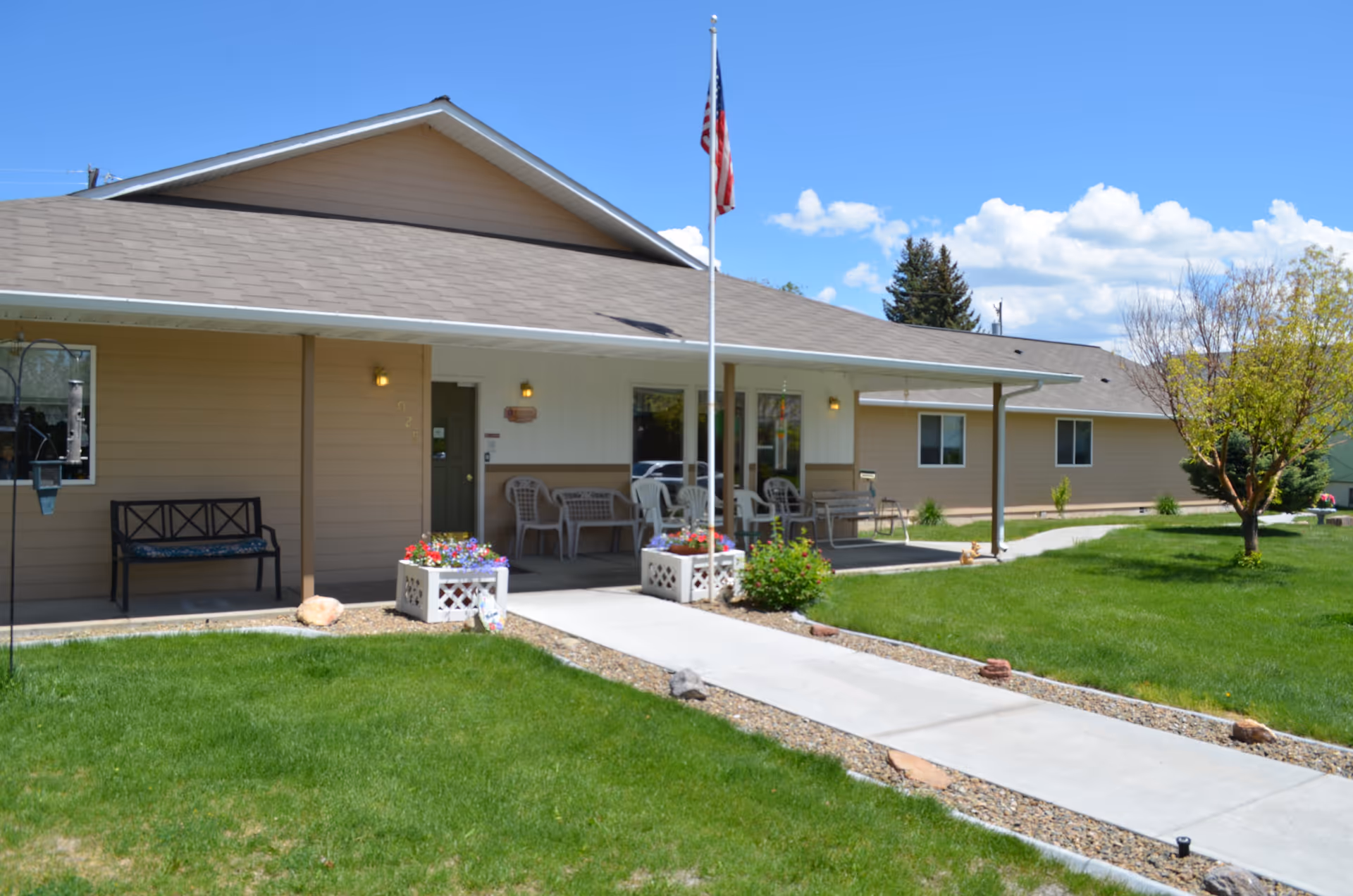 Exterior view of a single-story senior care facility building with beige siding and a gray roof. There is a concrete walkway leading to the entrance, which is shaded by a covered porch with several plastic chairs and benches. An American flag is mounted on a flagpole near the entrance. The surrounding area has green grass, flower planters, and a small tree under a blue sky with scattered clouds.