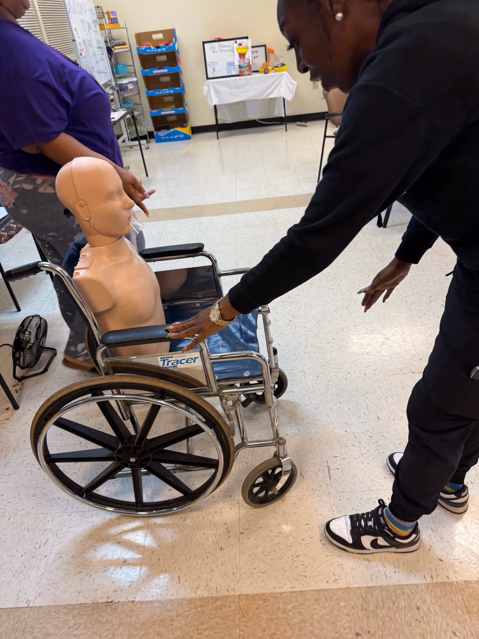 Caregiver interacts with a wheelchair-held medical mannequin in a facility training room.