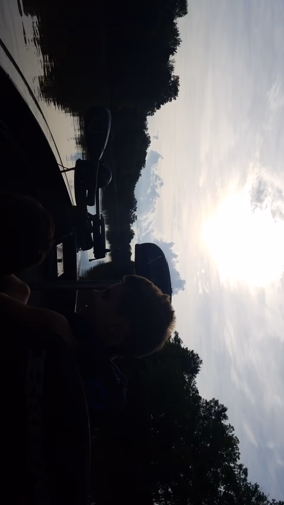 Silhouetted people beside a lake with boats and trees at sunset