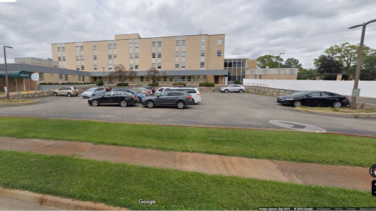 Exterior view of a multi-story brick facility with a parking lot and grassy foreground.