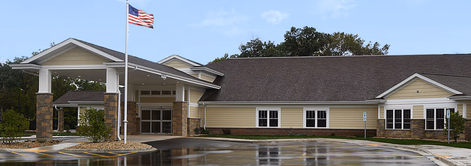 Front entrance of a single-story building with a covered porte-cochere, an American flag on a flagpole, and wet parking pavement reflecting the facade.