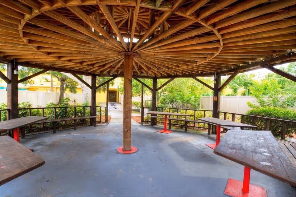 A wooden gazebo structure with a circular roof supported by a central wooden pillar and multiple beams. Inside the gazebo, there are several wooden picnic tables with red metal supports. Surrounding the gazebo are green bushes and trees, with a white fence and a yellow building visible in the background.
