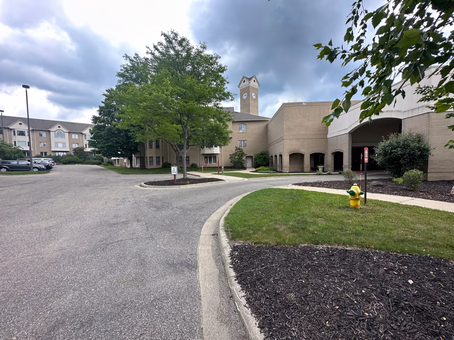 Exterior view of a senior living facility with a clock tower, surrounded by trees and landscaping. There is a parking area on the left side and a driveway leading to the building entrance on the right. The sky is cloudy.