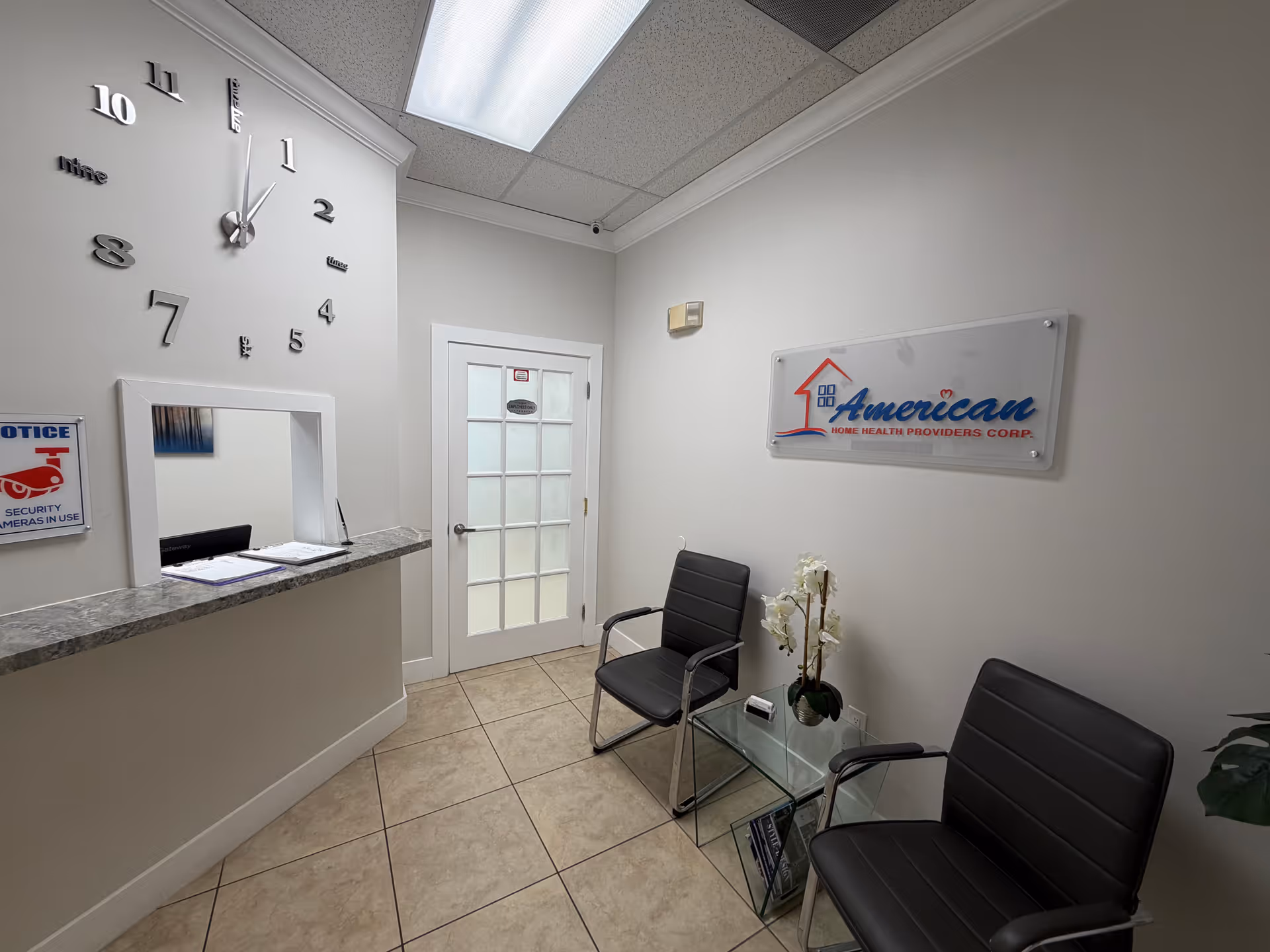 Reception area with a wall clock, counter, glass chairs, and American Home Health Providers Corp sign.