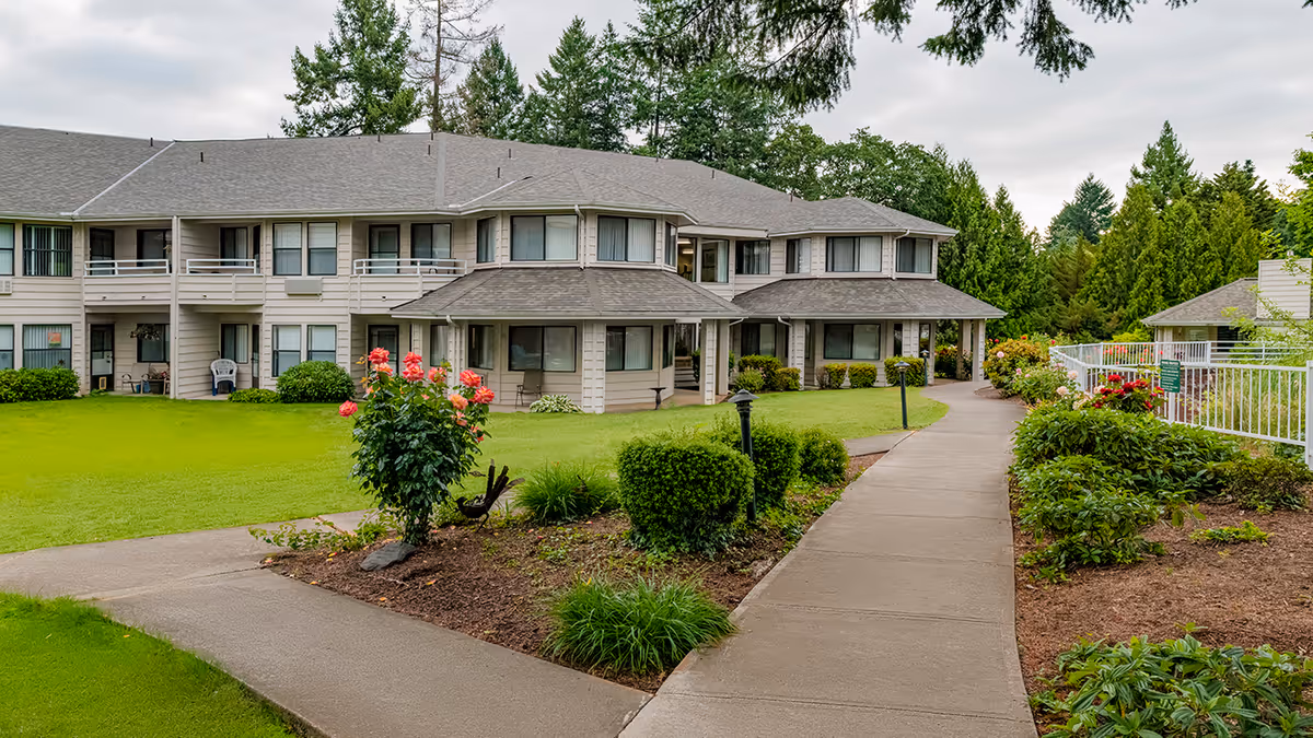Two-story senior living building with a paved walkway, landscaped lawn, and flowering bushes under a cloudy sky.
