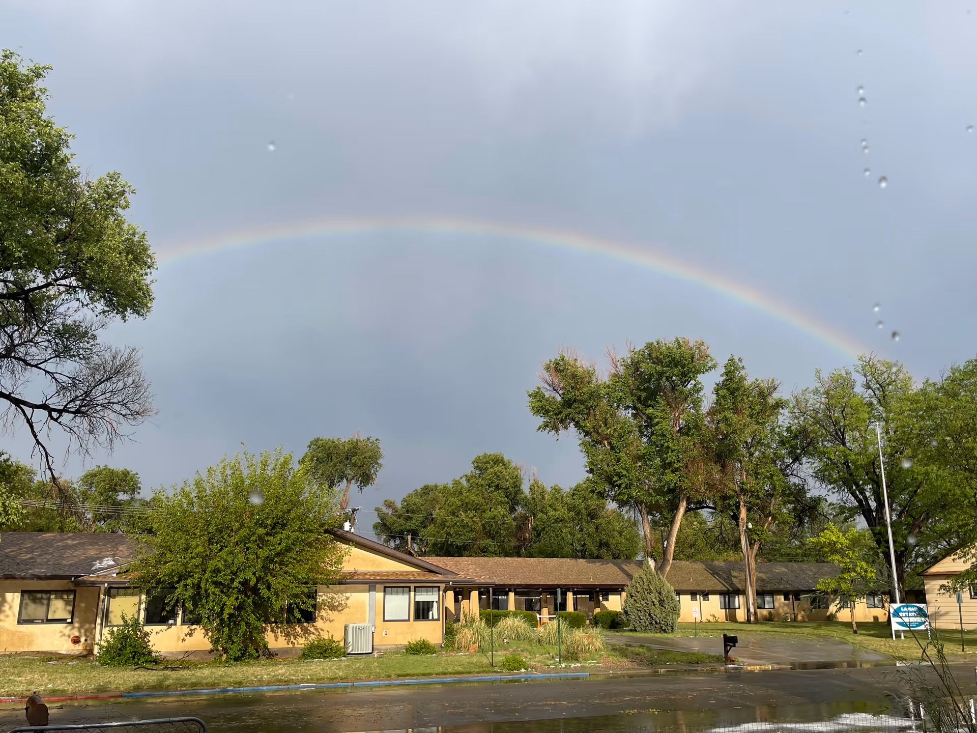 Rainbow arches over a single-story beige building with trees and a wet street.