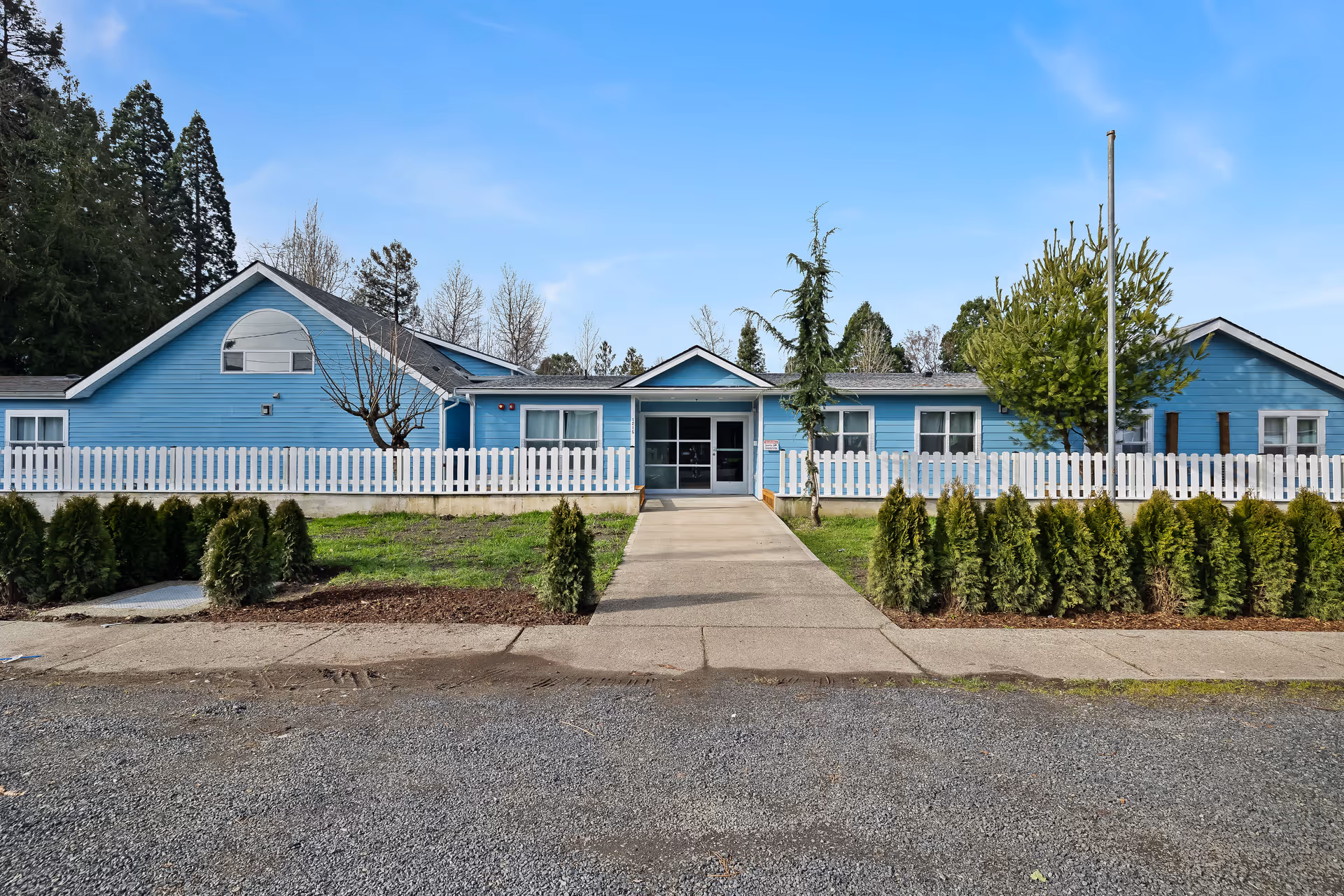 Front exterior of a single-story blue building with a white picket fence and a central walkway leading to the entrance.