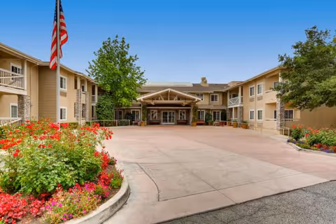 Exterior view of a senior living facility with a circular driveway, flowers, and flag.