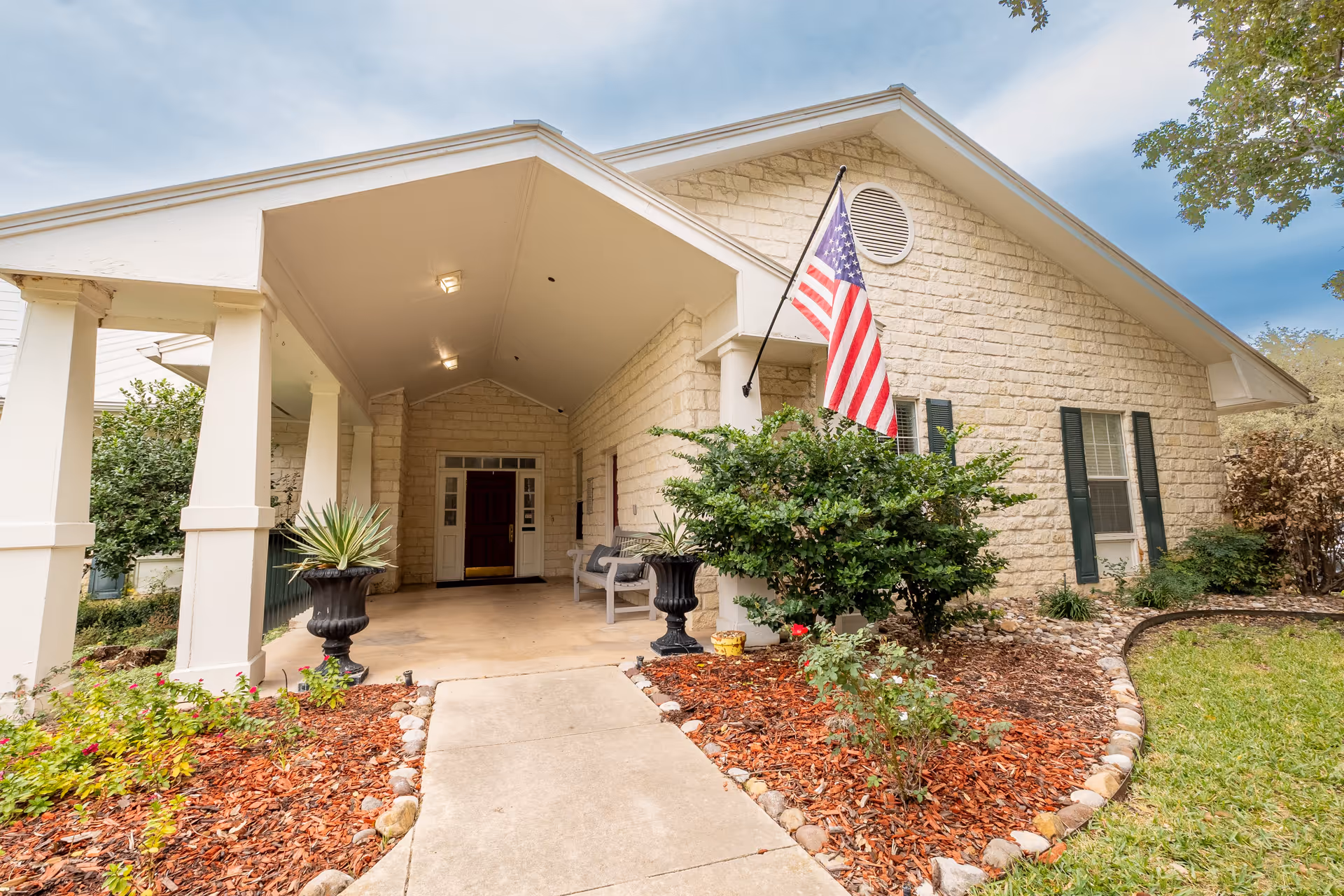 Front exterior view of a senior living facility with a covered entrance, stone facade, American flag, and landscaped garden beds with shrubs and flowers.