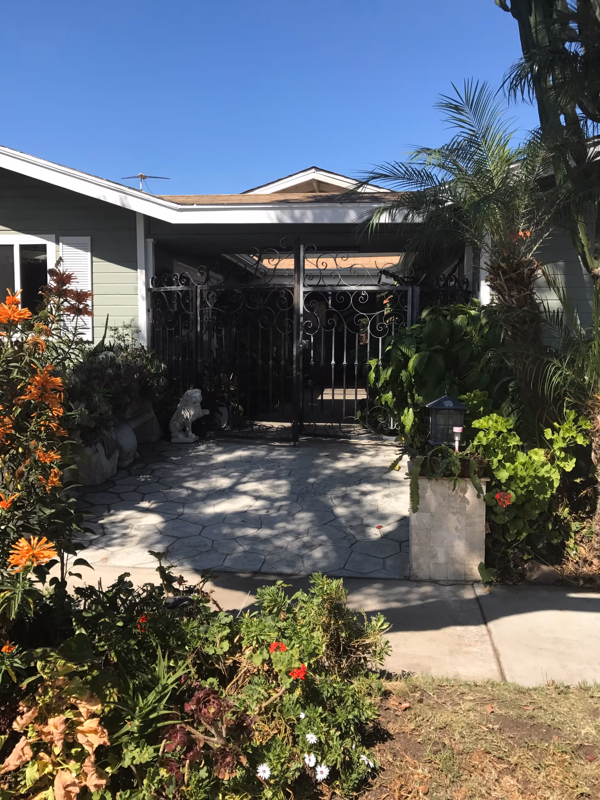Front entrance of a single-story home with a decorative iron gate, tiled walkway and lush landscaping under a clear blue sky.