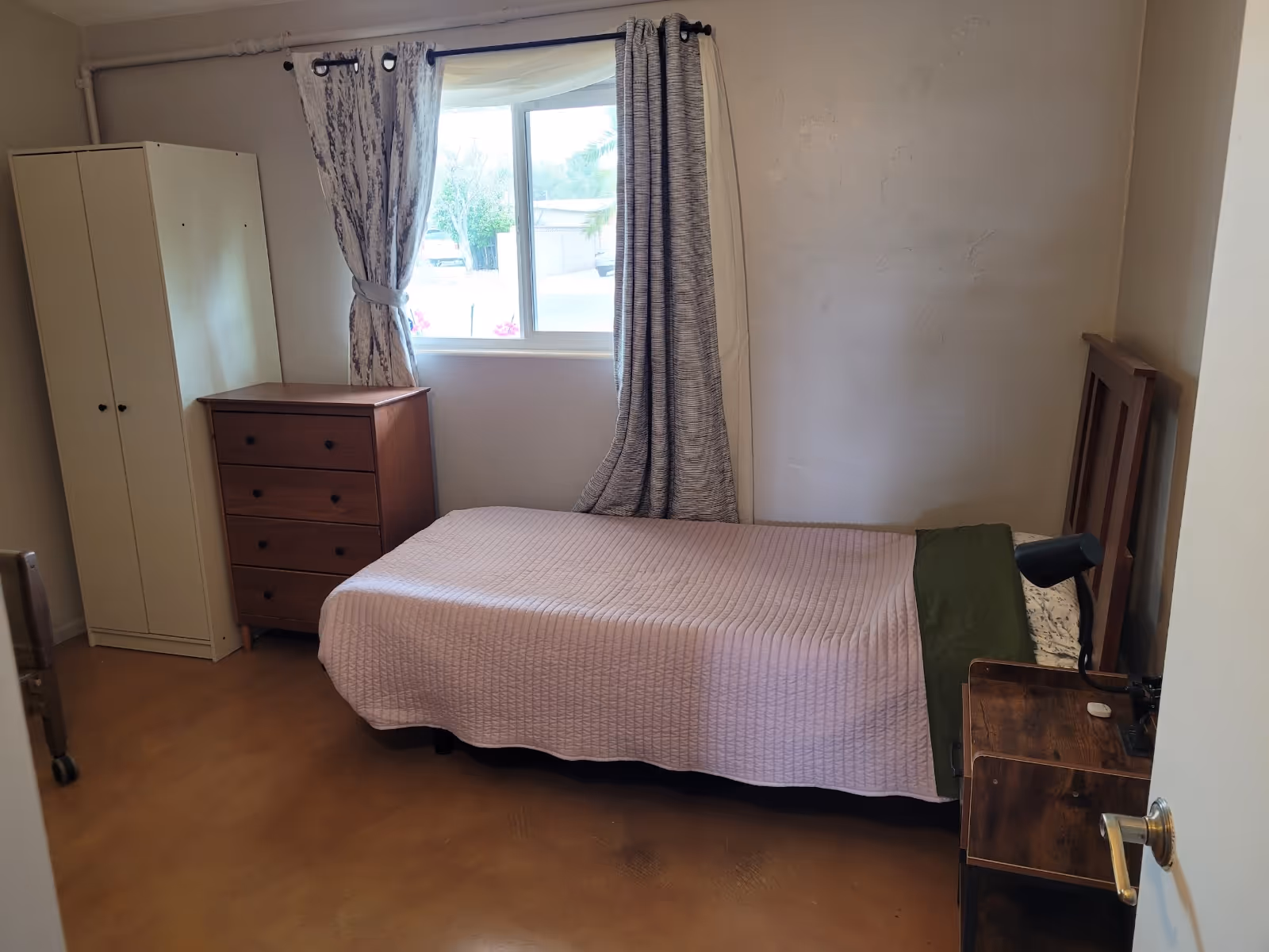 Small bedroom with single bed, dresser, wardrobe, nightstand lamp, and window with curtains.