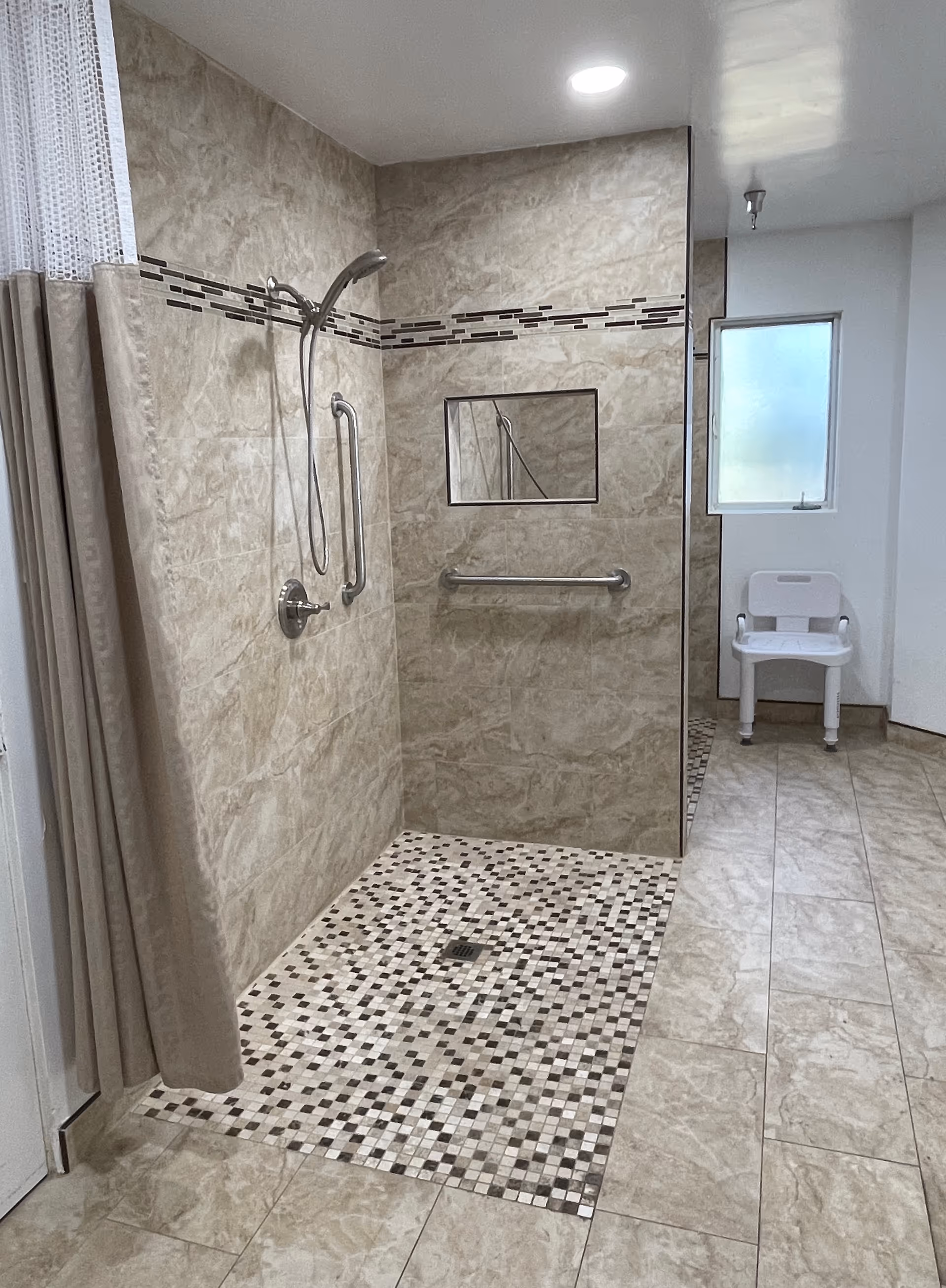 A spacious, accessible shower area with beige and brown tiled walls and floor. The shower has a handheld showerhead, grab bars, and a recessed shelf. A white plastic shower chair is positioned near a frosted window, and a beige shower curtain is partially visible on the left side.