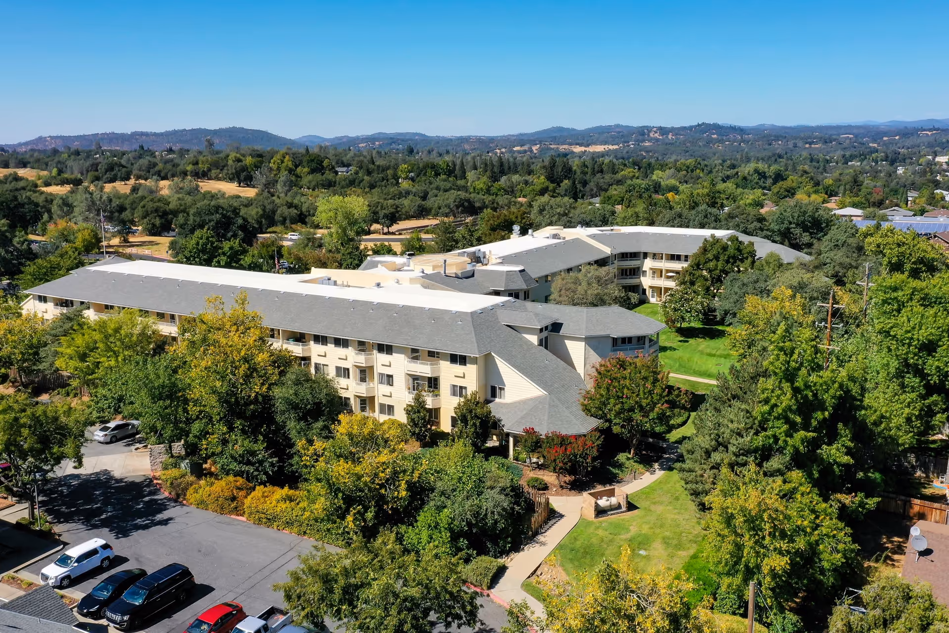 Aerial view of Solstice Senior Living At Auburn facility surrounded by trees and greenery, with a parking lot visible in the foreground and hills in the background under a clear blue sky.