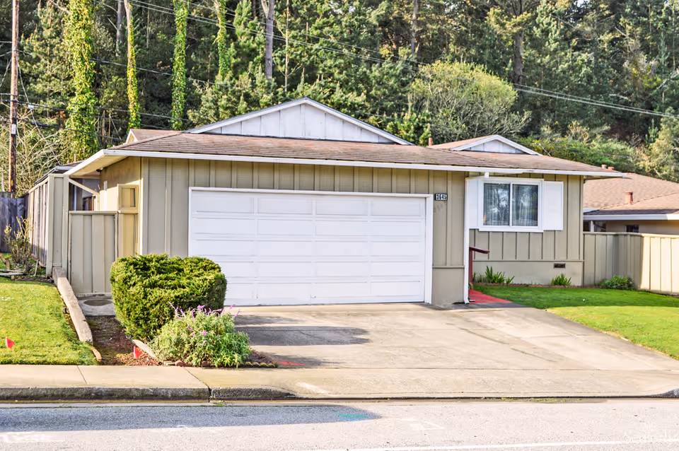 Front exterior view of a single-story house with a double garage door, a small front lawn with bushes and flowers, and a driveway leading to the garage. The house is surrounded by tall trees in the background.