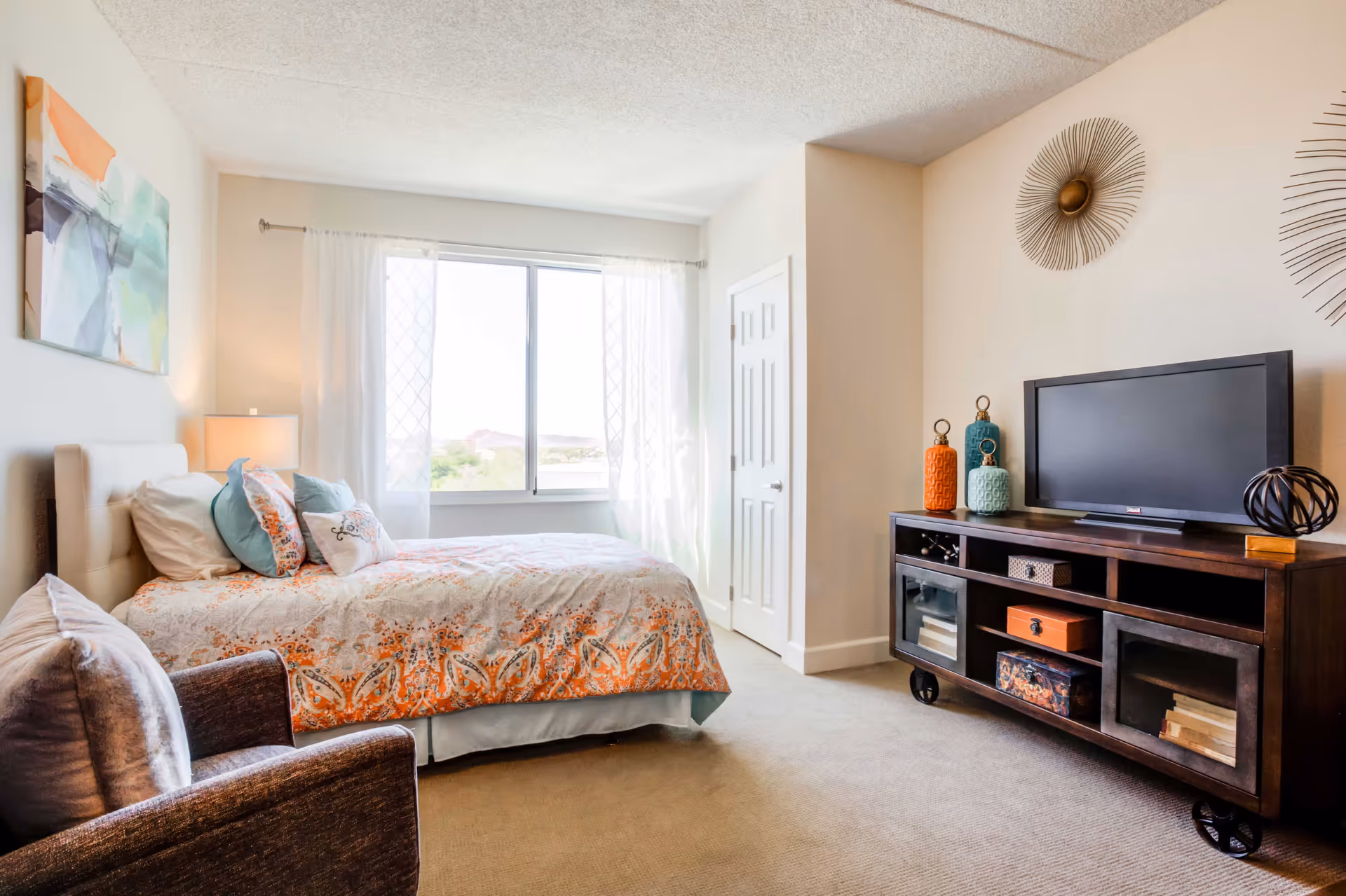 Sunlit bedroom with a bed dressed in patterned bedding, an armchair, and a TV on a wooden console beneath decorative wall art.