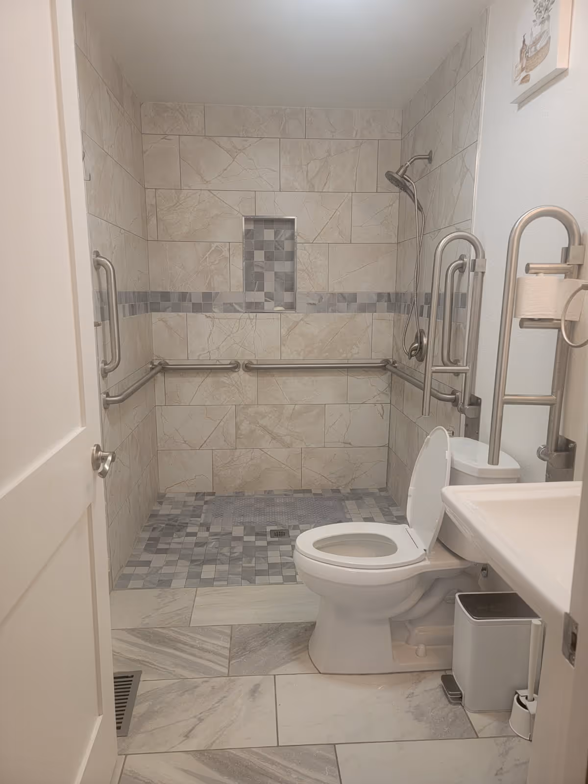 Accessible bathroom featuring tiled shower, multiple grab bars, toilet, and sink.
