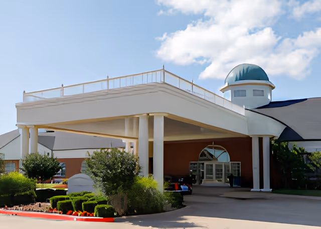 Front entrance of a senior living facility with a covered porte-cochère supported by columns, landscaped shrubs, and a domed cupola under a blue sky.