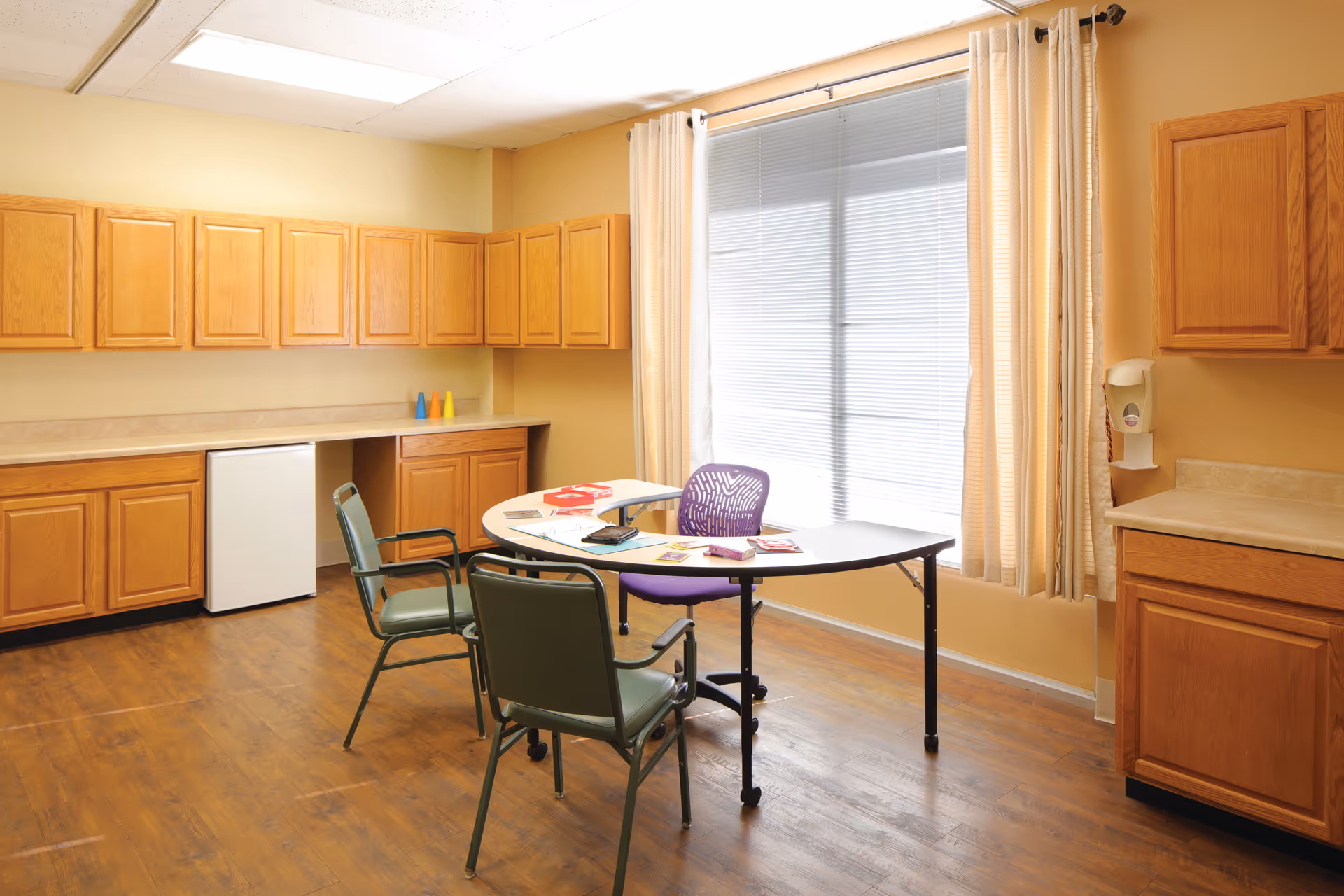 A small room with wooden cabinets along two walls, a mini refrigerator, and a curved table with three chairs. The table has some papers and books on it. A large window with closed blinds and beige curtains lets in natural light. The floor is wooden.