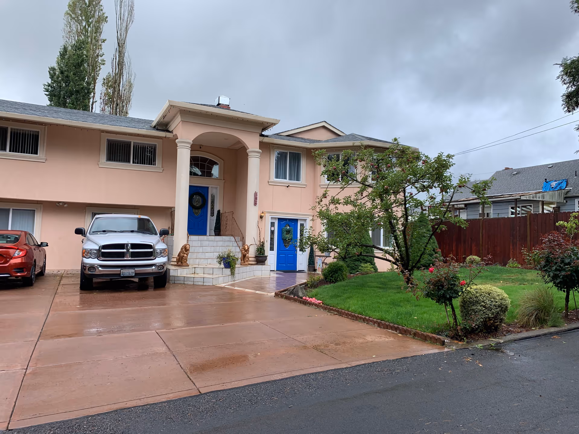 Exterior view of a two-story peach-colored building with two bright blue doors, a small staircase with two lion statues on either side, a driveway with two parked cars, and a green lawn with trees and bushes under a cloudy sky.