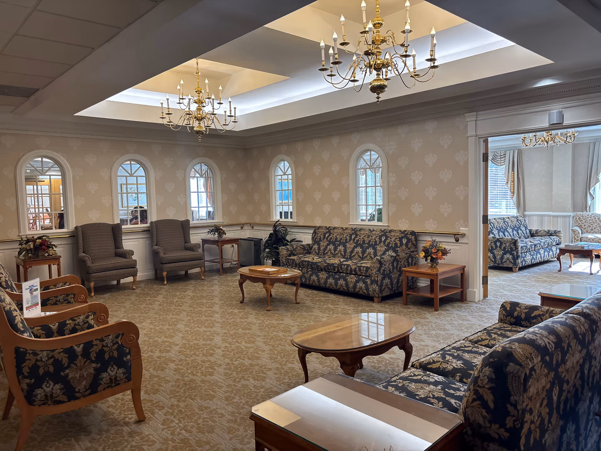 Elegant senior living lounge with chandeliers, arched windows, and blue floral seating.