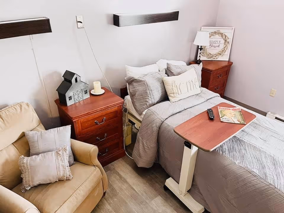 A cozy bedroom in a skilled nursing facility featuring a single bed with gray bedding and multiple pillows, a wooden bedside table with a lamp and decorative sign that reads 'Simply Blessed', a beige armchair with two pillows, and a small overbed table holding a remote control and a magazine.