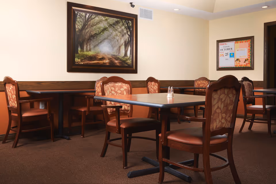 Empty dining area with wooden tables and upholstered chairs, a framed landscape painting and a bulletin board on the wall.