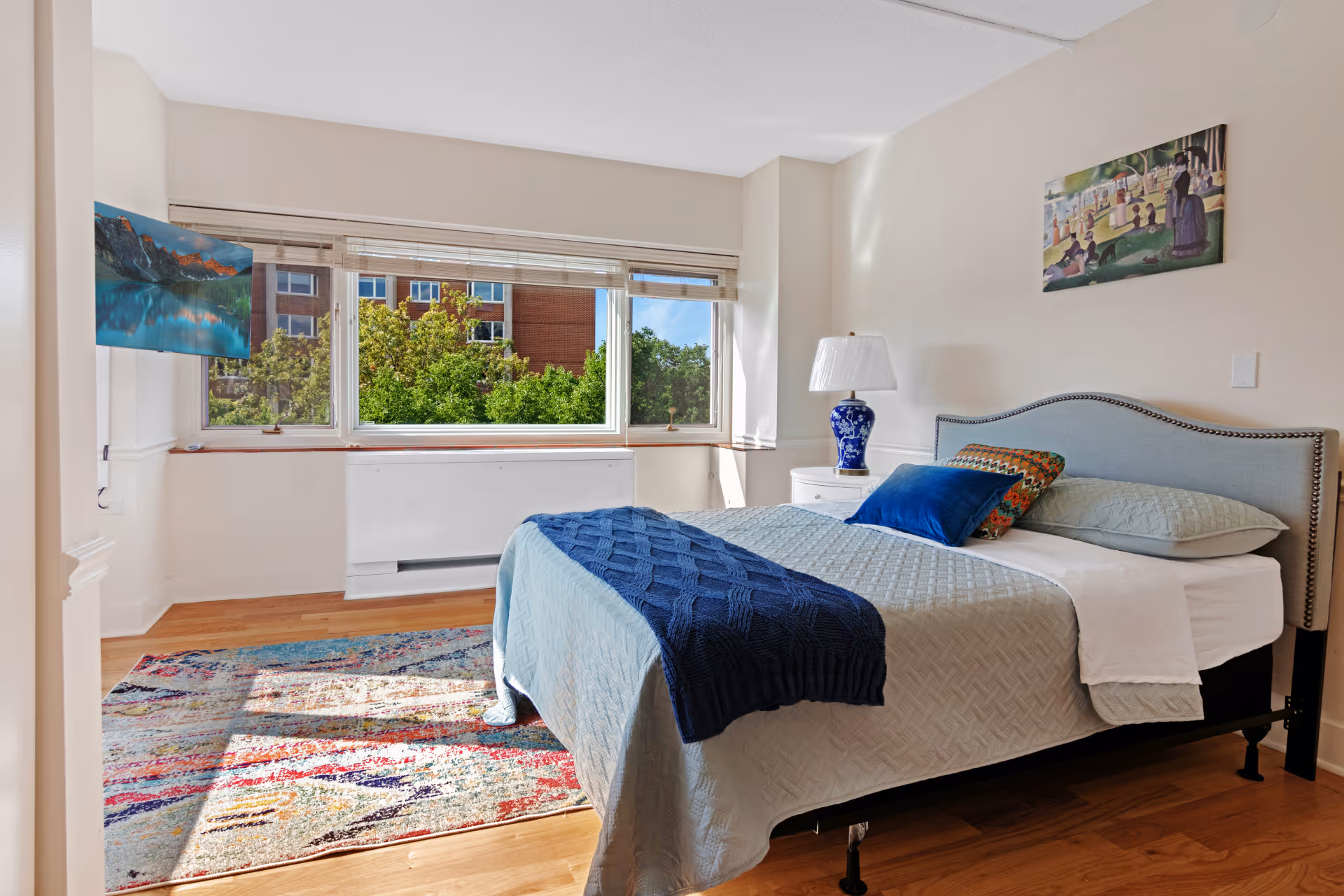 Sunny bedroom with a neatly made bed, bedside table and lamp, large window showing trees outside, and a colorful rug on hardwood floors.