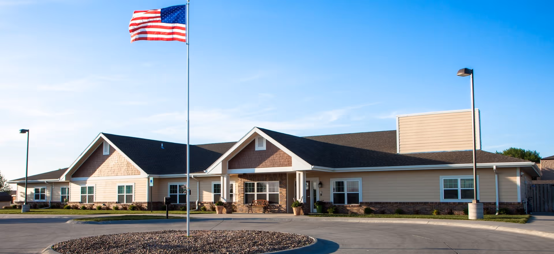 Single-story beige memory care building with a front entrance, circular driveway, and an American flag on a flagpole.