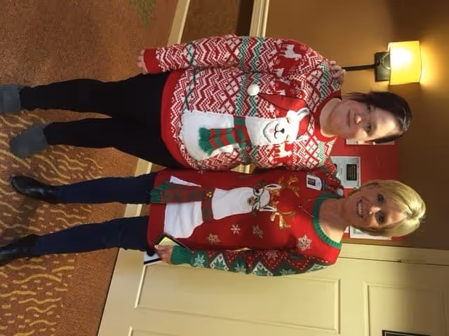 Two women standing indoors in a hallway, both wearing festive Christmas sweaters. One woman has a red sweater with a reindeer design, and the other has a red and white sweater with a polar bear wearing a scarf. The hallway has patterned carpet, beige walls, a door, and a wall-mounted lamp.