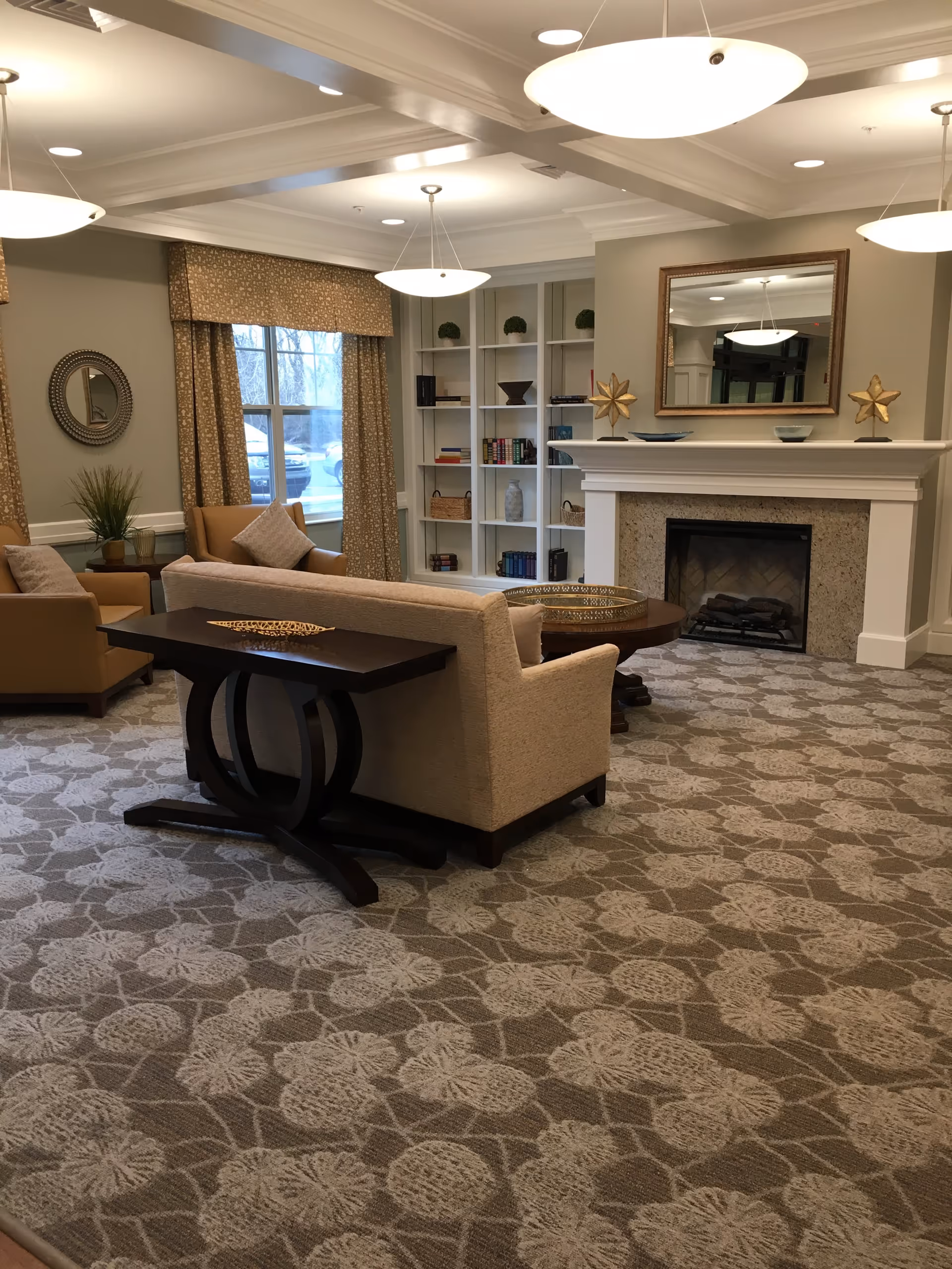 A cozy senior living lounge with sofas and armchairs arranged around a fireplace, built-in bookshelves, and overhead pendant lights.