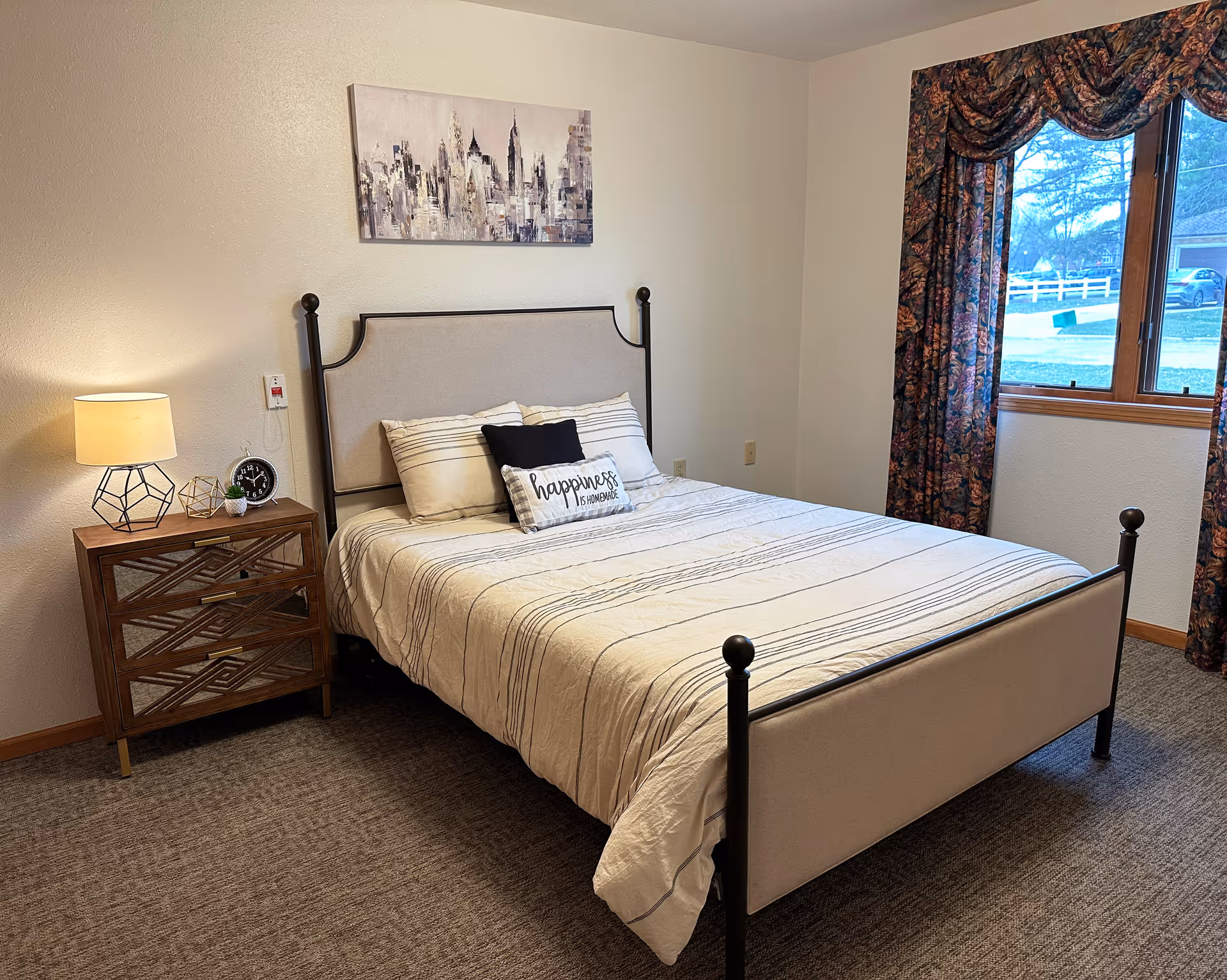 A neatly made bed with a beige upholstered headboard and footboard in a bedroom. The bed has striped bedding and three pillows, one of which has the text 'happiness is homemade'. Next to the bed is a wooden nightstand with a lamp, a clock, and decorative items. A window with floral curtains shows an outdoor view with trees and a fence. A painting of a city skyline hangs on the wall above the bed.
