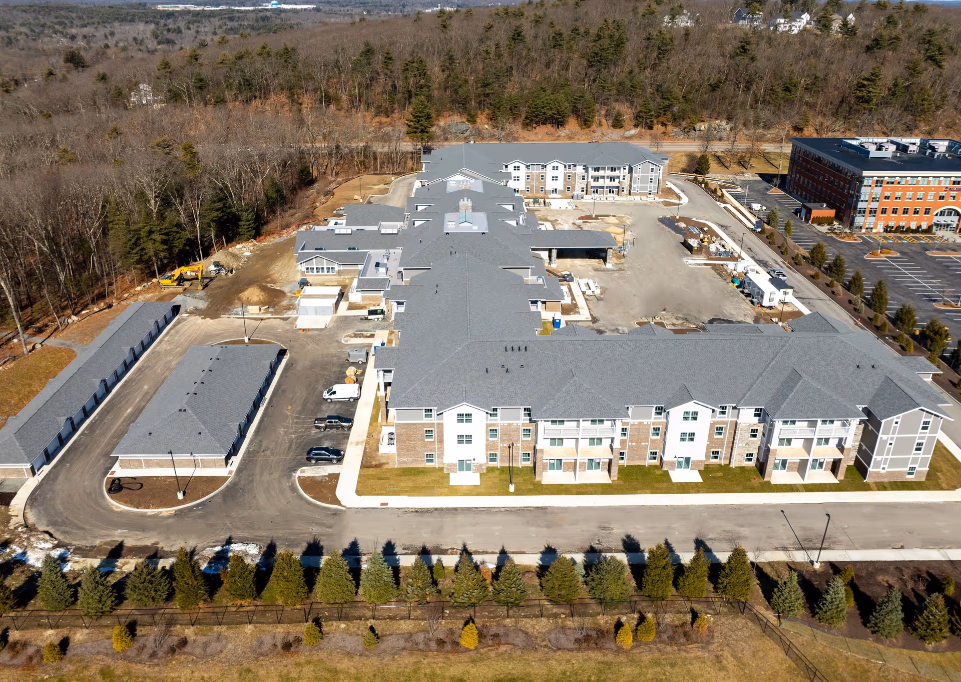 Aerial view of Pleasant Hill Retirement Community showing a large multi-story building with gray roofs, surrounded by parking areas, trees, and some construction equipment. The building is situated near a wooded area and other buildings are visible in the background.