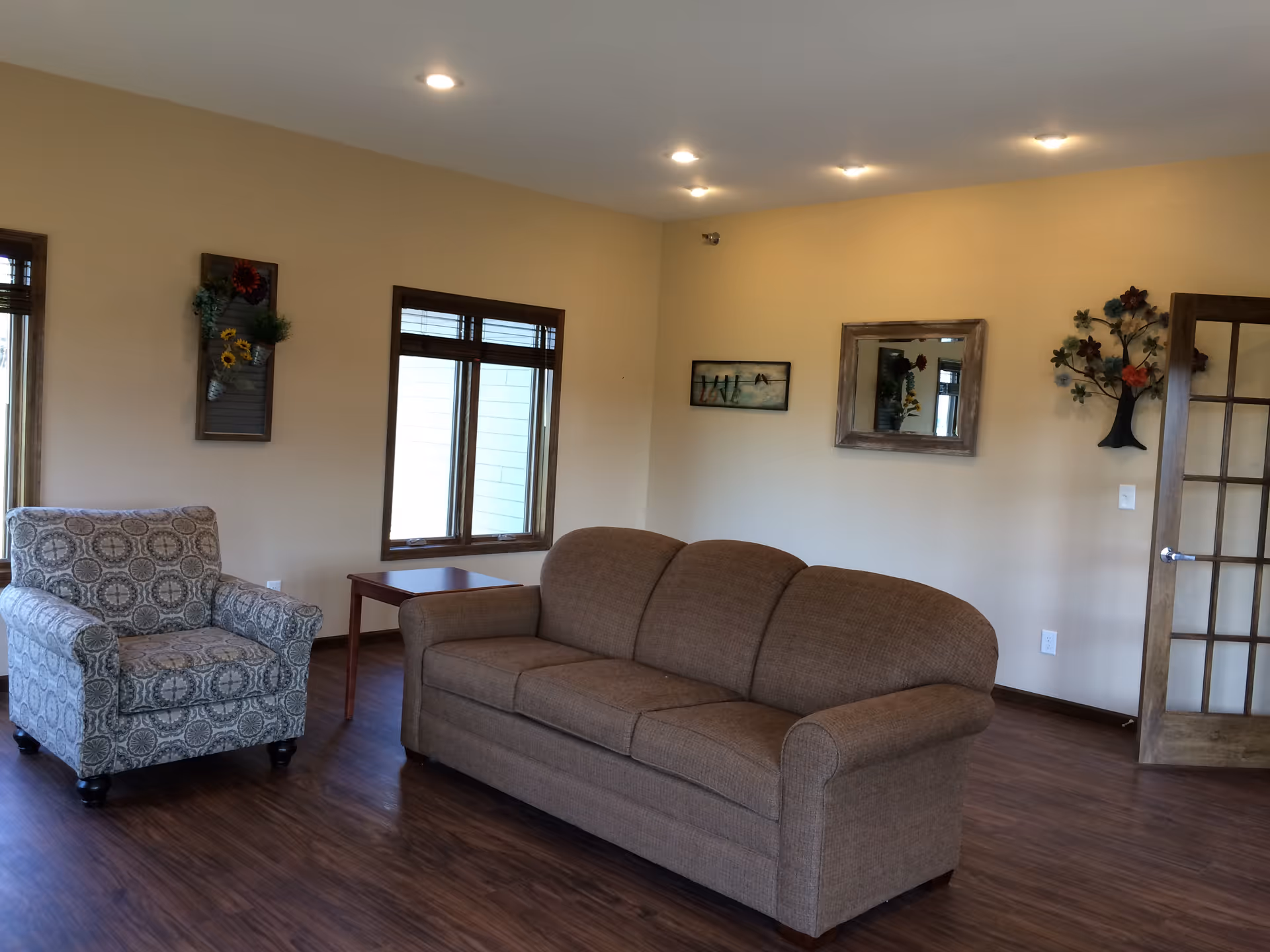 Spacious living room with a brown sofa, patterned armchair, wood floors, windows and wall decorations.