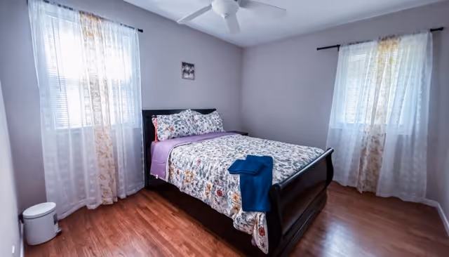 A cozy bedroom with a dark wood bed, floral quilt, sheer curtains, and wooden floor.