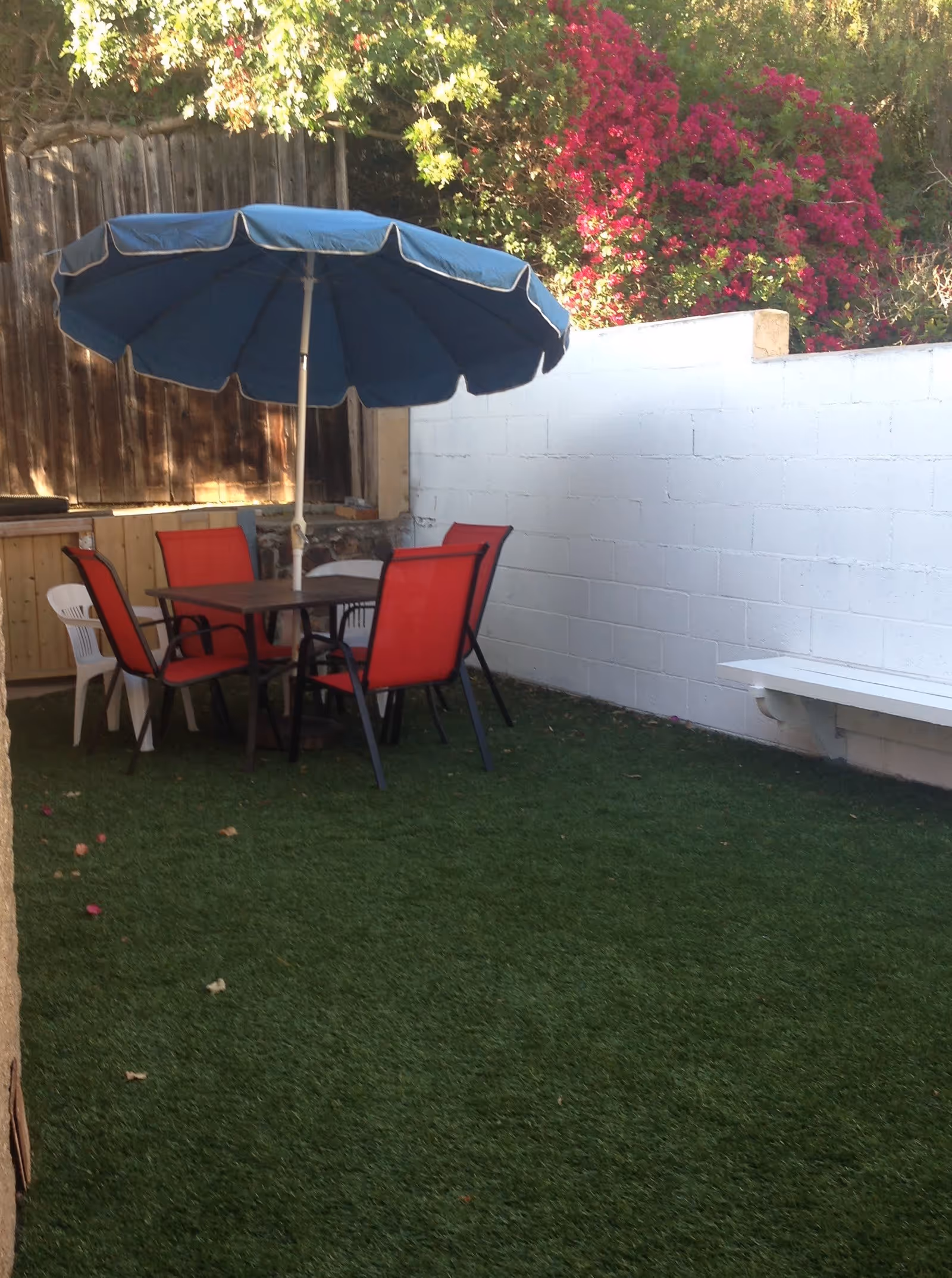 Outdoor patio area with artificial grass, a table with a blue umbrella, four red chairs, two white chairs, a white bench attached to a white brick wall, and vibrant pink flowers growing over the wall.